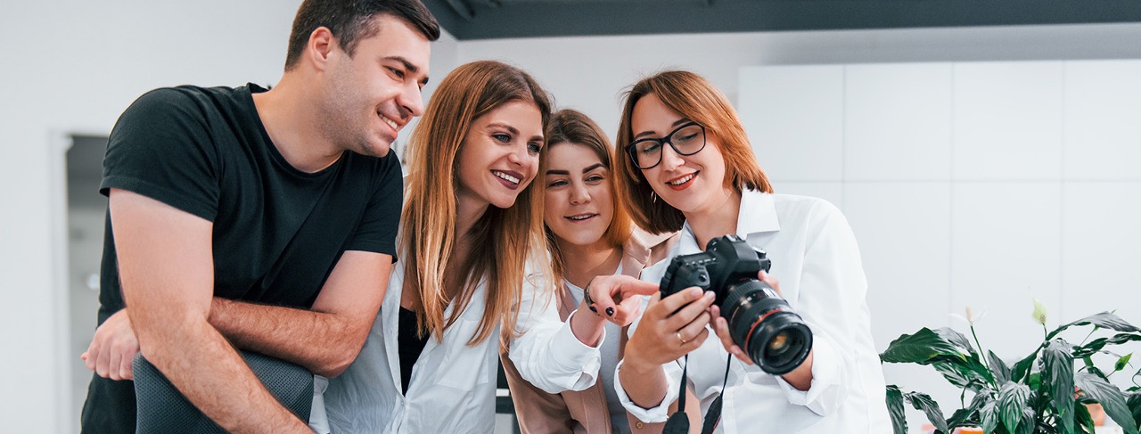 Czwórka młodych ludzi, elegancko ubranych pochyla sie nad aparatem fotograficznym. Tło białe, widać w prawym dolnym rogu roślinę.