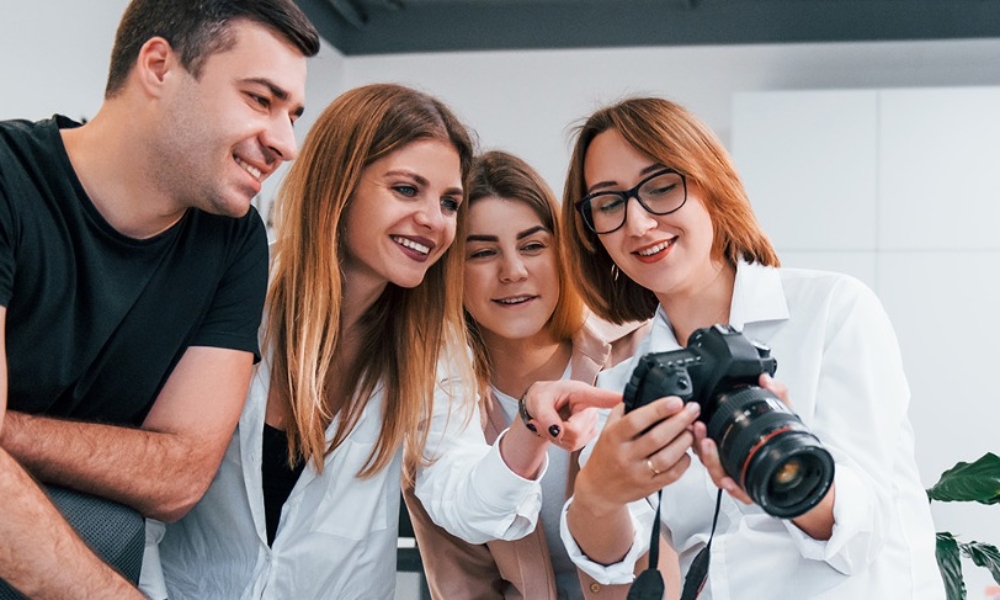 Czwórka młodych ludzi, elegancko ubranych pochyla sie nad aparatem fotograficznym. Tło białe, widać w prawym dolnym rogu roślinę.